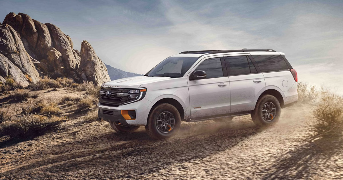 White Ford Expedition SUV driving on dusty desert trail with rocky mountains in background