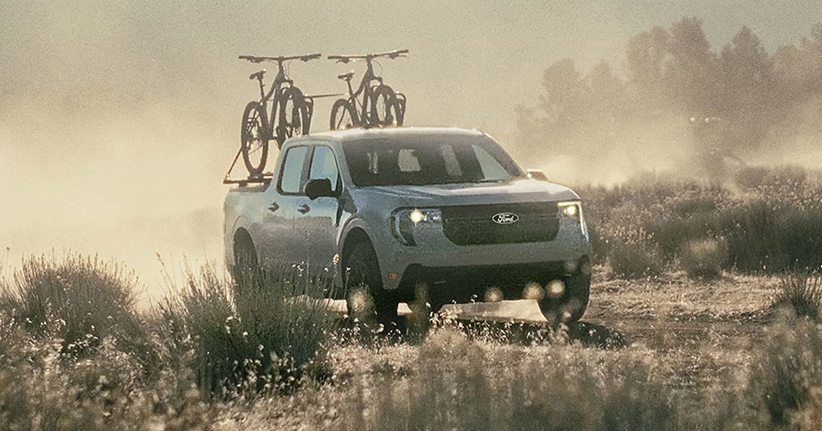 Ford Maverick pickup truck with bicycles on roof rack, driving through misty grassy field at sunrise