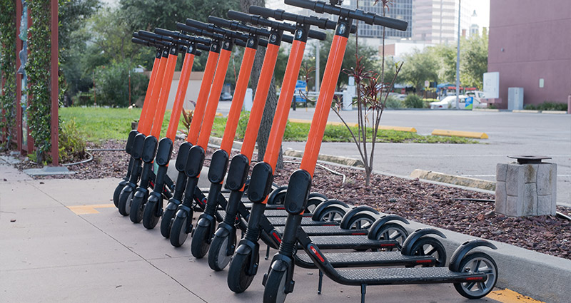 A line of dockless electric scooters, with orange handlebars and black bodies, are neatly parked side-by-side on a sidewalk in an urban area. In the background, there are trees, a parking lot, and the faint outline of city buildings. The scooters appear to be from the Spin company, judging by the visible color scheme.