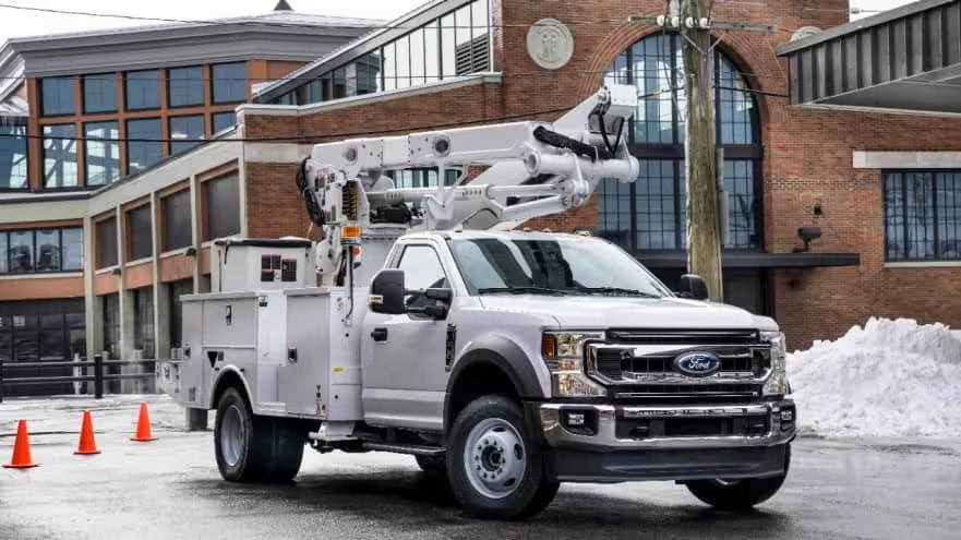 White Ford F-550 utility truck with bucket lift boom parked on snowy street near brick building with traffic cones